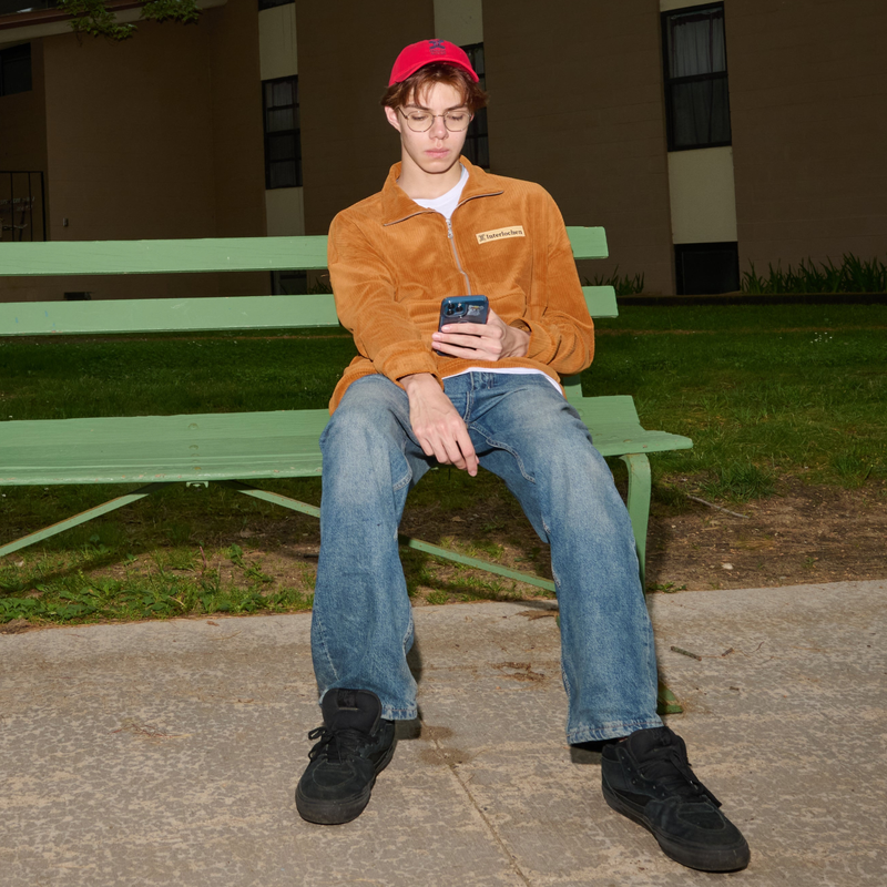 Person sitting on a green bench using a phone, wearing a brown jacket and red cap.
