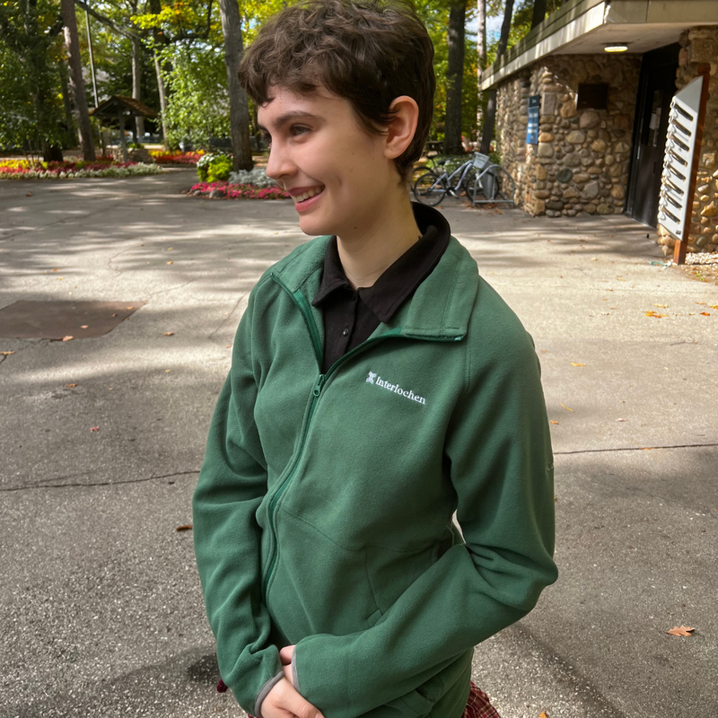Person wearing a green Interlochen jacket standing outdoors with trees and a building in the background.