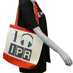 A red canvas tote bag with 'I LISTEN TO IPR' printed in black and a graphic of headphones on the front, with red trim, displayed on a mannequin.