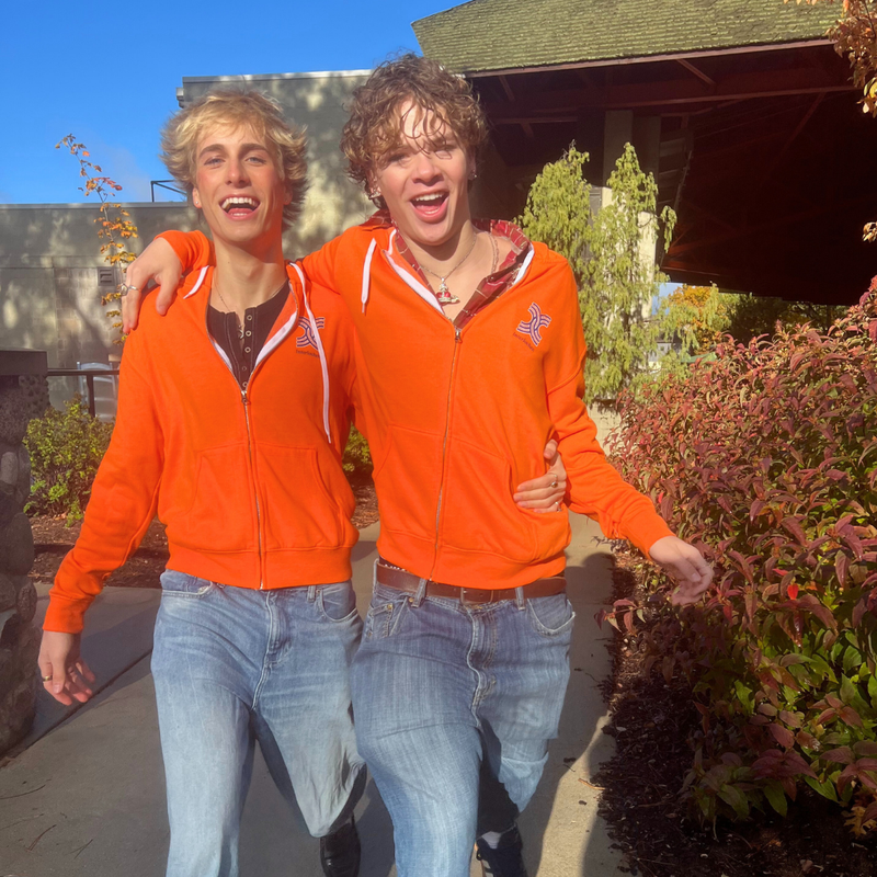 Two people wearing orange hoodies and jeans standing outdoors.