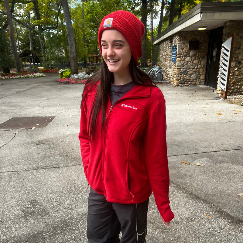 Person wearing a red  columbia jacket and beanie standing outdoors near a stone building.
